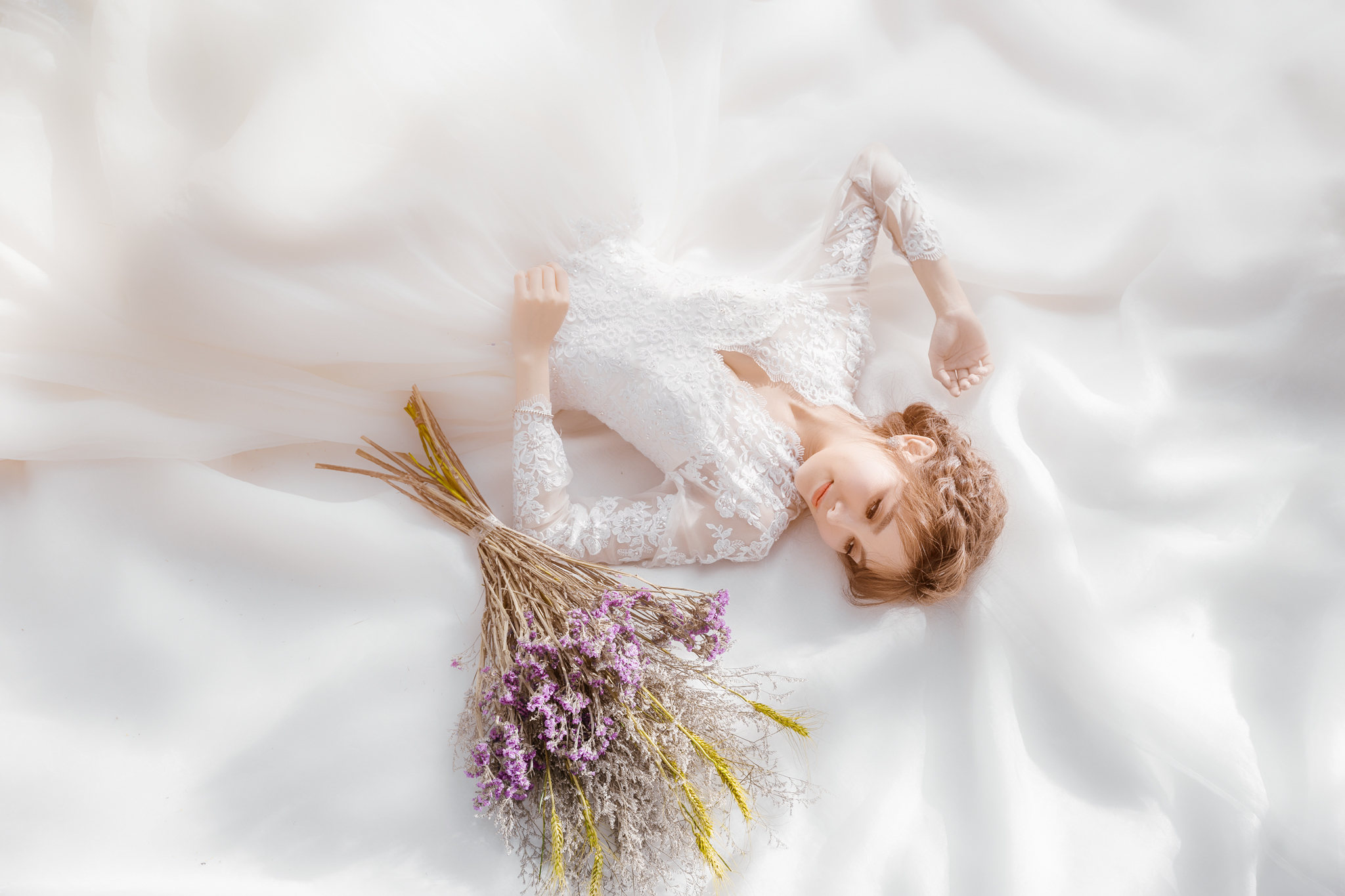 Noiva elegante deitada em tecido branco segurando buquê de flores